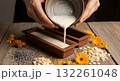 A close-up shot of a person pouring natural oils from small glass beakers into a creamy white soap base in a wooden bowl, then stirring the mixture with a wooden spoon to blend the ingredients 132261048