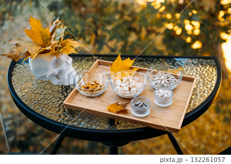 Outdoor glass table setup featuring a skull adorned with autumn leaves next to a wooden tray filled with assorted supplements. Mortality and health awareness in autumn visuals, memento mori, Outdoor glass table setup featuring a skull adorned with autumn leaves next to a wooden tray filled with assorted supplements. Mortality and health awareness in autumn visuals, memento mori, 132261057