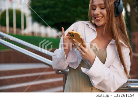 Smiling, in headphones and with smartphone. Beautiful female student is outdoors near the university Smiling, in headphones and with smartphone. Beautiful female student is outdoors near the university 132261154