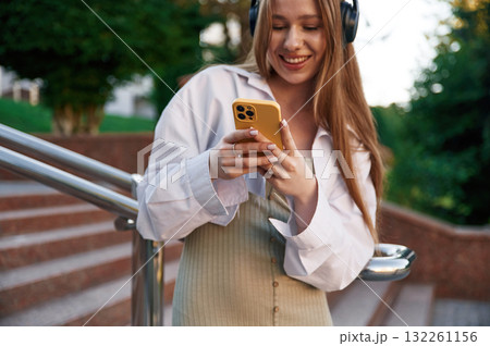 Smiling, in headphones and with smartphone. Beautiful female student is outdoors near the university Smiling, in headphones and with smartphone. Beautiful female student is outdoors near the university 132261156