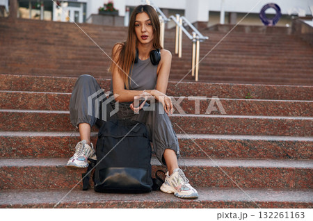 Holding smartphone, sitting on the stairs. Beautiful female student is outdoors near the university 132261163