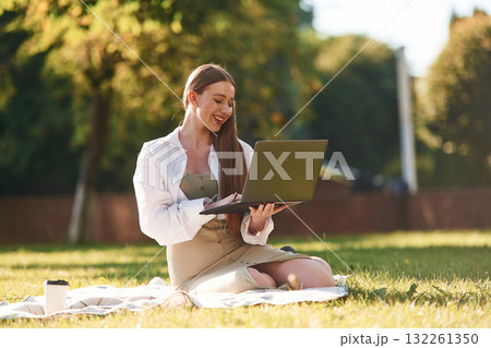 On the grass with laptop. Beautiful female student is outdoors near the university On the grass with laptop. Beautiful female student is outdoors near the university 132261350