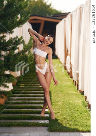 Happy young woman in bikini is standing outdoors near the fence and fir tree Happy young woman in bikini is standing outdoors near the fence and fir tree 132261614