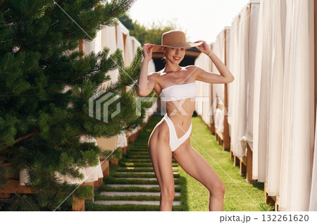In the hat. Happy young woman in bikini is standing outdoors near the fence and fir tree In the hat. Happy young woman in bikini is standing outdoors near the fence and fir tree 132261620