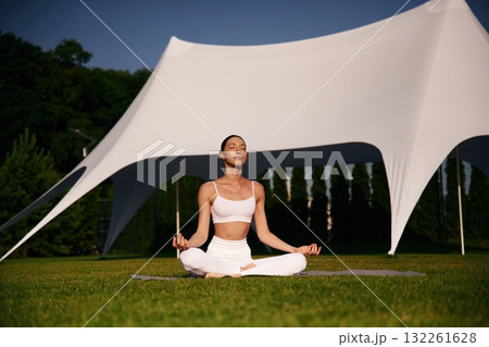 Sitting in the lotus pose. Attractive young fitness woman is on the green grass at daytime Sitting in the lotus pose. Attractive young fitness woman is on the green grass at daytime 132261628