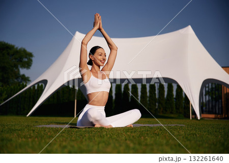 White tent behind, doing yoga. Attractive young fitness woman is on the green grass at daytime 132261640