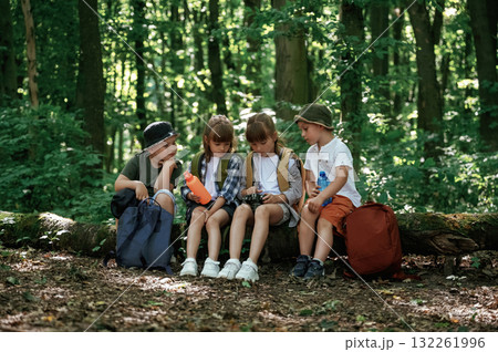 Taking a rest, holding camera and water bottle. Kids in forest at summer daytime together Taking a rest, holding camera and water bottle. Kids in forest at summer daytime together 132261996