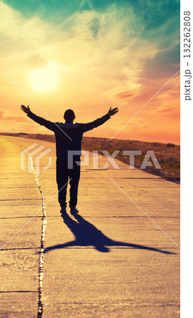 A young man with hands in the air walks along a concrete road and looks at a beautiful orange sunset. Vertical image A young man with hands in the air walks along a concrete road and looks at a beautiful orange sunset. Vertical image 132262808