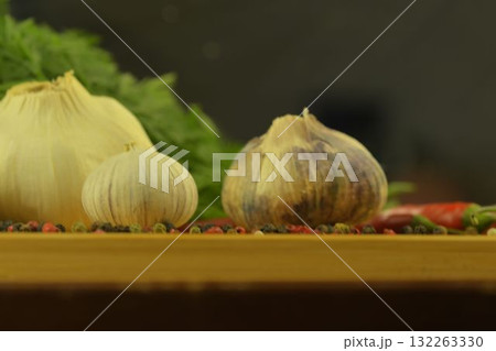 A bulb of garlic, red chili peppers, four-color pepper, and carrot tops on a wooden cutting board. The concept of culinary arts and vegetables in the home. Macro image, close-up. Garlic - species A bulb of garlic, red chili peppers, four-color pepper, and carrot tops on a wooden cutting board. The concept of culinary arts and vegetables in the home. Macro image, close-up. Garlic - species 132263330