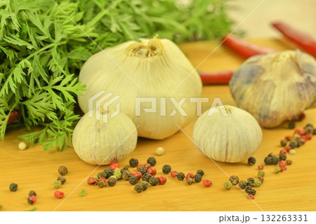 A bulb of garlic, red chili peppers, four-color pepper, and carrot tops on a wooden cutting board. The concept of culinary arts and vegetables in the home. Macro image, close-up. Garlic - species A bulb of garlic, red chili peppers, four-color pepper, and carrot tops on a wooden cutting board. The concept of culinary arts and vegetables in the home. Macro image, close-up. Garlic - species 132263331