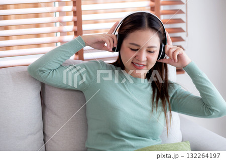 Happy young asian woman wearing headphones listening music while sitting on sofa with fun. 132264197
