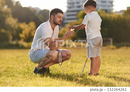 With badminton rackets. Father and little son are together on the summer field With badminton rackets. Father and little son are together on the summer field 132264261