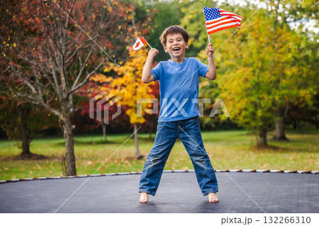 Smiling boy jumping high on trampoline outdoors holding Canada and USA flags in his hands on a bright autumn day Smiling boy jumping high on trampoline outdoors holding Canada and USA flags in his hands on a bright autumn day 132266310