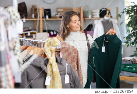 Positive young girl standing in interior of clothing boutique and choosing dress for autumn-winter season Positive young girl standing in interior of clothing boutique and choosing dress for autumn-winter season 132267854