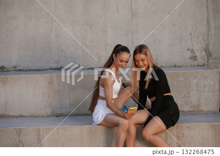 Holding smartphone, taking a break. Two sportive women are outdoors together Holding smartphone, taking a break. Two sportive women are outdoors together 132268475