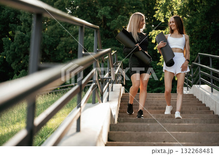 Carrying yoga mats. Two sportive women are outdoors together Carrying yoga mats. Two sportive women are outdoors together 132268481