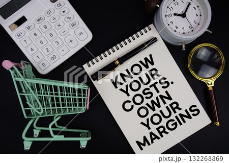Know your costs Own your margins text written on notepad with alarm clock and trolley shopping cart on black background 132268869