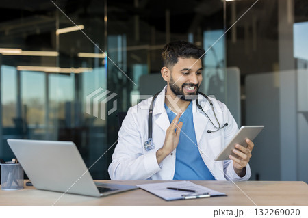 Happy male doctor in uniform and stethoscope making a video call on a digital tablet. Offering remote medical consultation and online healthcare services from a modern office with a laptop on the desk 132269020