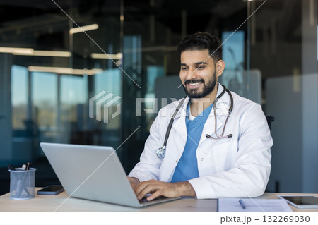 Male indian doctor smiling at laptop while providing telehealth consultation from office desk, confident and professional during virtual patient care and remote medical support Male indian doctor smiling at laptop while providing telehealth consultation from office desk, confident and professional during virtual patient care and remote medical support 132269030