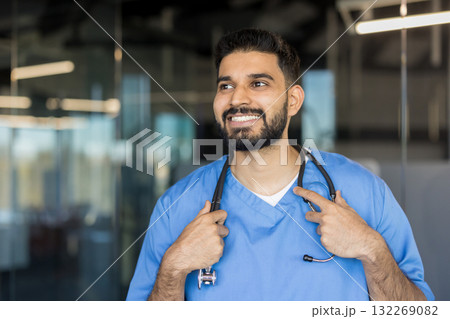 Young indian male medical professional wearing blue scrubs and a stethoscope around his neck, looking away and smiling confidently in a modern hospital or clinic setting 132269082