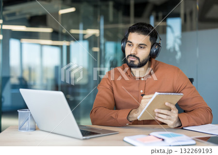 Young man wearing headphones and using a laptop for online learning or a webinar, intently focusing on a virtual lecture and diligently jotting down important notes in a modern office Young man wearing headphones and using a laptop for online learning or a webinar, intently focusing on a virtual lecture and diligently jotting down important notes in a modern office 132269193