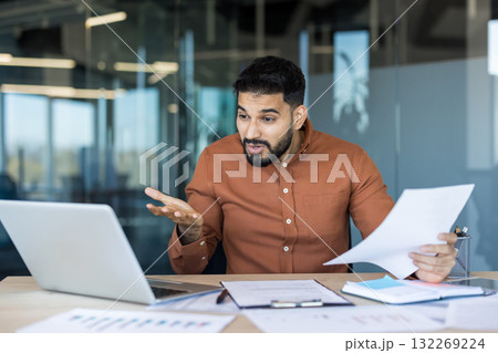 Frustrated young businessman confused by difficult work, looking at a laptop and holding paper documents while sitting at an office desk, illustrating concepts of stress and workload Frustrated young businessman confused by difficult work, looking at a laptop and holding paper documents while sitting at an office desk, illustrating concepts of stress and workload 132269224