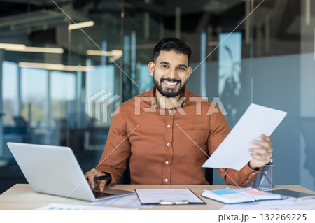Smiling indian businessman sitting at office desk, reviewing financial documents and using a laptop, representing success, productivity, confidence, and modern business Smiling indian businessman sitting at office desk, reviewing financial documents and using a laptop, representing success, productivity, confidence, and modern business 132269225