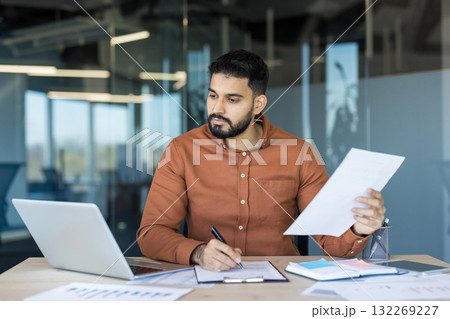 Young man working at a desk, reviewing documents and signing papers while using a laptop in a contemporary office environment, symbolizing business management and professional productivity Young man working at a desk, reviewing documents and signing papers while using a laptop in a contemporary office environment, symbolizing business management and professional productivity 132269227