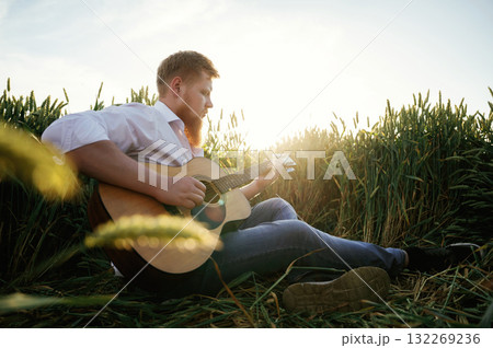 Guitar in hands. Man in white shirt is on the agricultural field Guitar in hands. Man in white shirt is on the agricultural field 132269236