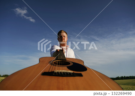 Holding acoustic guitar. Man in white shirt is on the agricultural field 132269490