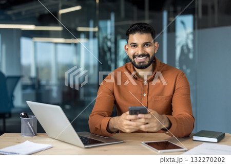 Smiling indian businessman sitting at an office desk, holding a smartphone while working with a laptop and tablet, portraying connectivity and professionalism 132270202