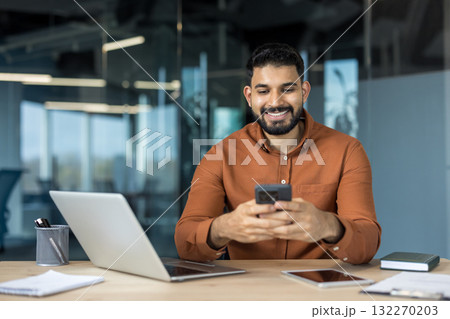 Young indian businessman sitting at his desk in a contemporary office, happily navigating on his smartphone, representing concepts like mobile communication, business technology, and connectivity 132270203