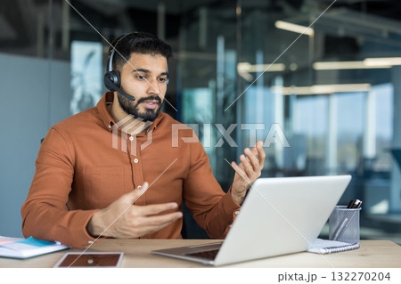 Young bearded man in a brown shirt working in a modern office, having a video conference call on a laptop, actively communicating and gesturing with hands 132270204