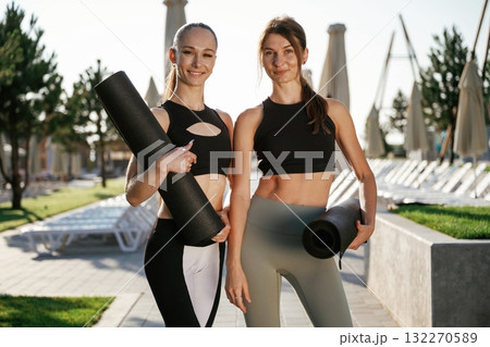 Black yoga mats in hands, standing. Two fitness women are together outdoors, conception of sport Black yoga mats in hands, standing. Two fitness women are together outdoors, conception of sport 132270589