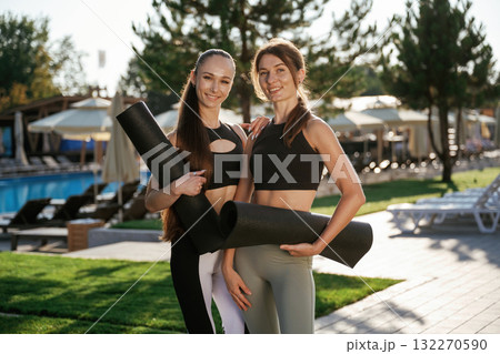 Black yoga mats in hands, standing. Two fitness women are together outdoors, conception of sport Black yoga mats in hands, standing. Two fitness women are together outdoors, conception of sport 132270590