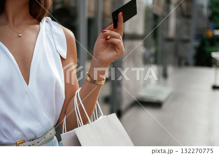 Black credit card in hand. Stylish woman is with shopping bag outdoors near the building Black credit card in hand. Stylish woman is with shopping bag outdoors near the building 132270775