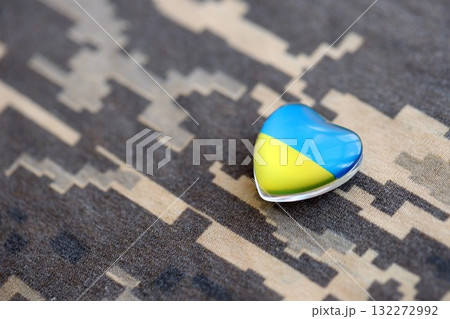 Glass heart with Ukrainian flag lies on military camouflage fabric. Concept of support and defend Ukraine Glass heart with Ukrainian flag lies on military camouflage fabric. Concept of support and defend Ukraine 132272992