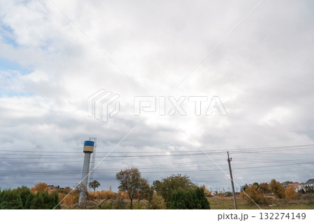 Rural Landscape with Silver Water Tower under Cloudy Autumn Sky. Trees, Power Lines, and Distant Houses. Rural Landscape with Silver Water Tower under Cloudy Autumn Sky. Trees, Power Lines, and Distant Houses. 132274149