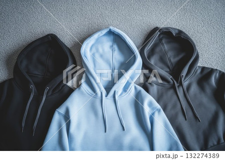 Black, gray, and blue hoodies neatly arranged on smooth gray surface. Casual fashion displayed in natural lighting. Neural network AI generated 132274389