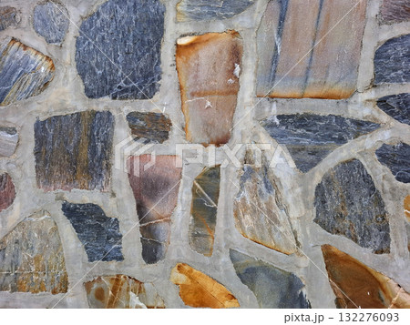 This stone wall features a unique blend of multi-colored natural stones, creating a rich texture. Sunlight enhances the warm hues of gray, orange, and brown, highlighting the craftsmanship. 132276093