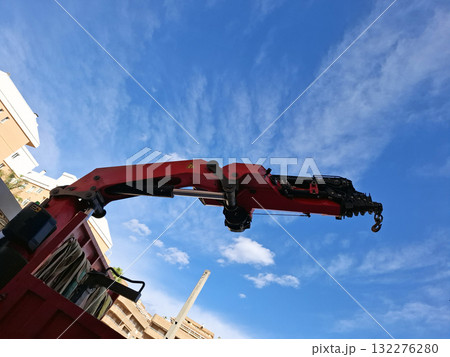 A truck mounted crane extends its telescopic boom high against a clear blue sky filled with clouds. The scene is set in an urban area, showcasing construction activity nearby. 132276280
