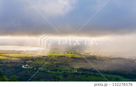 A sweeping valley landscape with green fields, winding roads, and misty sunrays 132276576