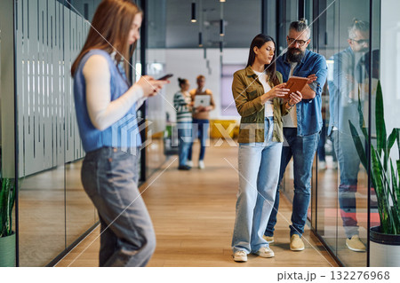 Two business professionals having a focused discussion while reviewing information on a tablet in a bright modern office hallway, symbolizing teamwork, communication, and digital collaboration. Two business professionals having a focused discussion while reviewing information on a tablet in a bright modern office hallway, symbolizing teamwork, communication, and digital collaboration. 132276968