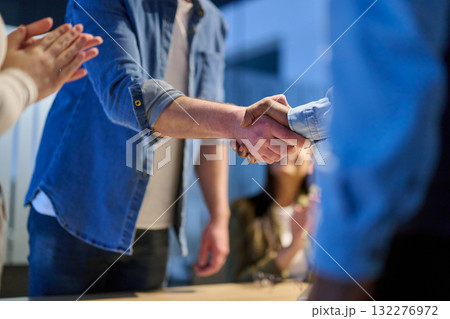 Two businessmen shaking hands after successful negotiation in a modern office, symbolizing partnership, agreement, success, and teamwork. A woman in the background applauds the deal. 132276972