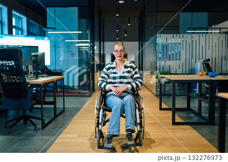 A confident smiling business woman in a wheelchair sitting in a bright modern office hallway, representing workplace inclusion, accessibility, and professional success. 132276973