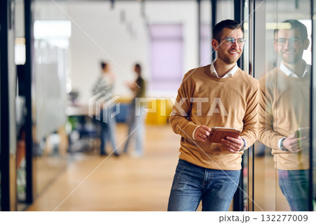 Businessman standing by glass wall in a modern office corridor, holding a digital tablet and smiling with reflection visible on the glass, symbolizing success, optimism, and modern business lifestyle. Businessman standing by glass wall in a modern office corridor, holding a digital tablet and smiling with reflection visible on the glass, symbolizing success, optimism, and modern business lifestyle. 132277009
