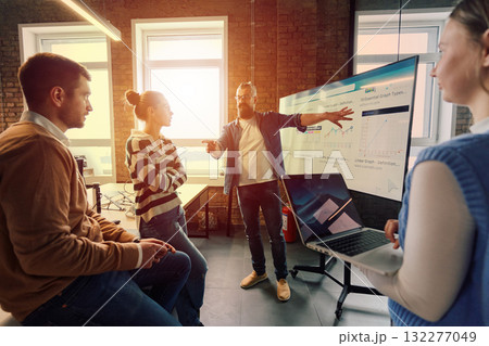 Group of professionals gathered in a modern office for a business presentation. A man with a beard explains data charts on a large screen while the team listens attentively. Concept of teamwork 132277049