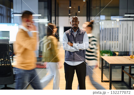 Confident african american businessman standing with crossed arms in the center of a busy office hallway while colleagues move around him, symbolizing leadership, focus, and stability in a dynamic 132277072