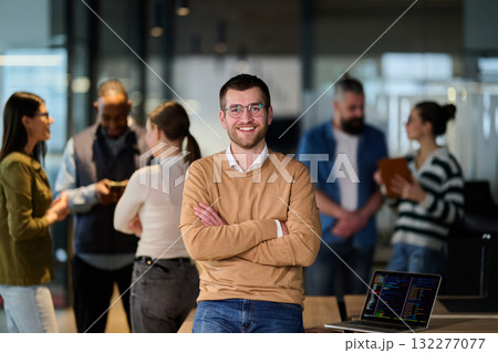 Portrait of a confident young businessman wearing glasses and a beige sweater, standing with arms crossed in a modern office while colleagues collaborate in the background. 132277077