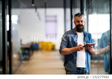 Bearded businessman standing in modern office hallway, holding digital tablet and looking confidently at camera, representing experience, leadership, and innovation in contemporary business 132277082